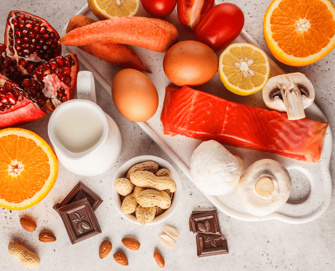 Assorted foods on a table: salmon, eggs, milk, oranges, pomegranate, tomatoes, chocolate, peanuts, carrots, and mushrooms.