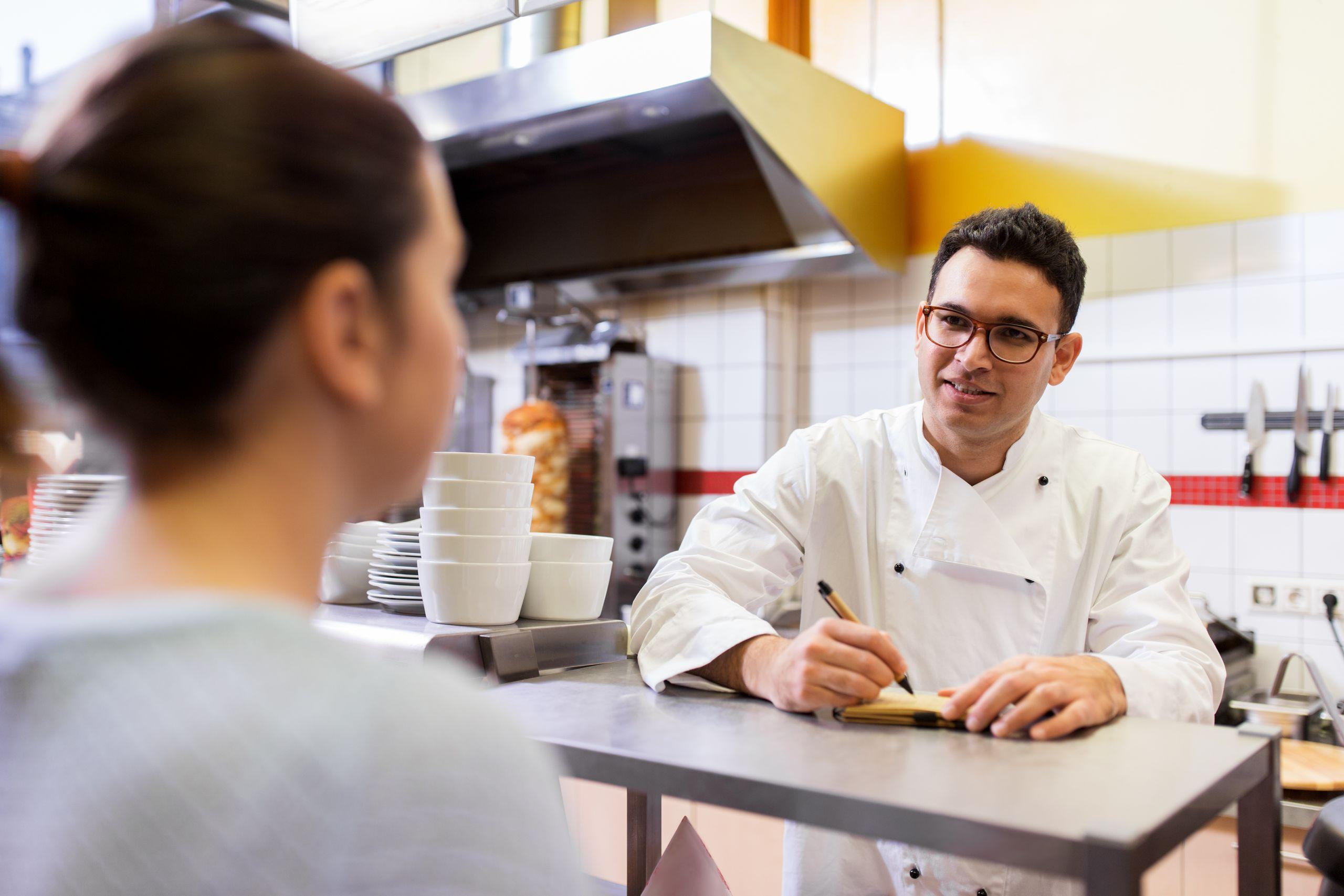 Chef taking an order from a customer at a counter in a restaurant kitchen, with bowls and kitchen equipment in the background.