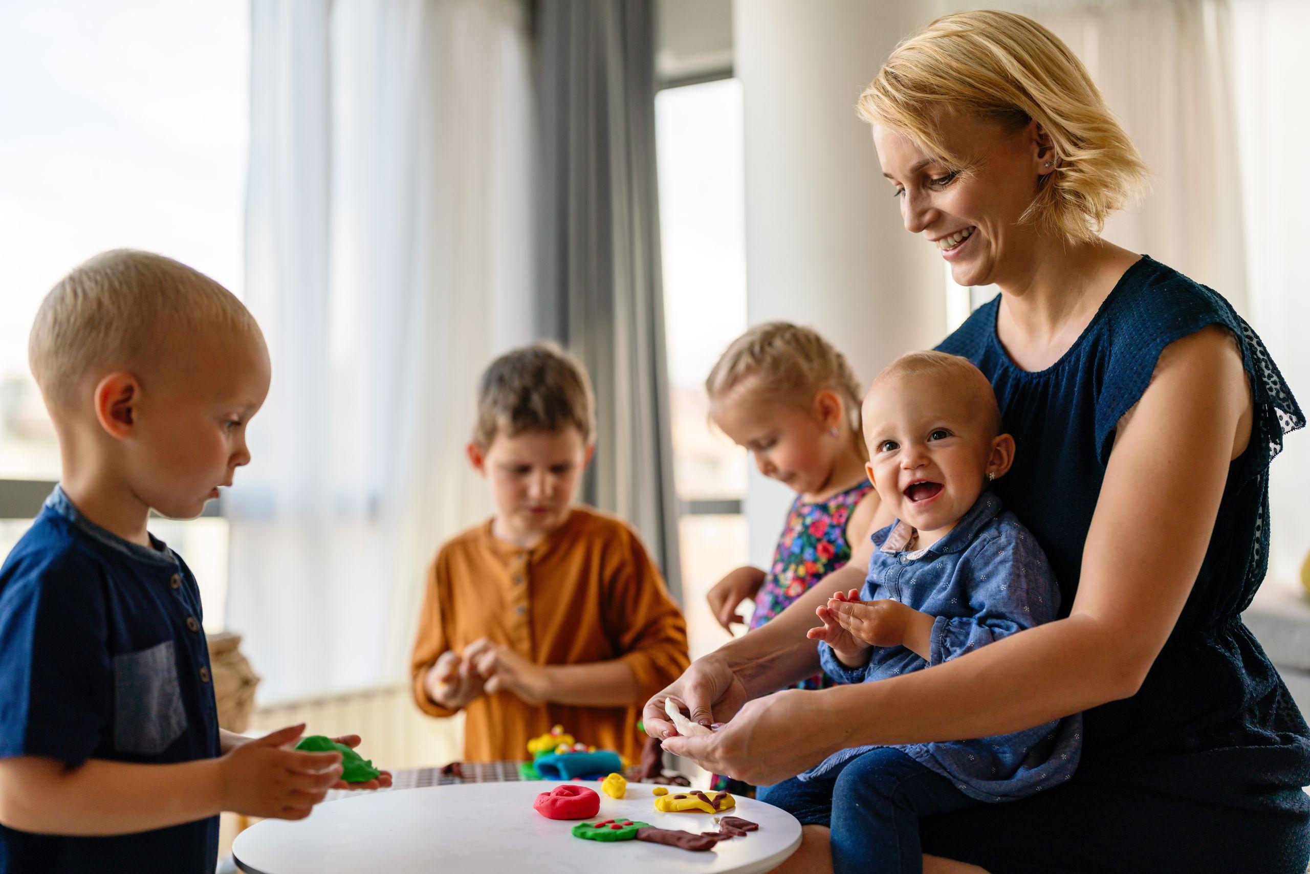 A woman and four children are playing with colorful clay at a white table in a bright room with large windows.