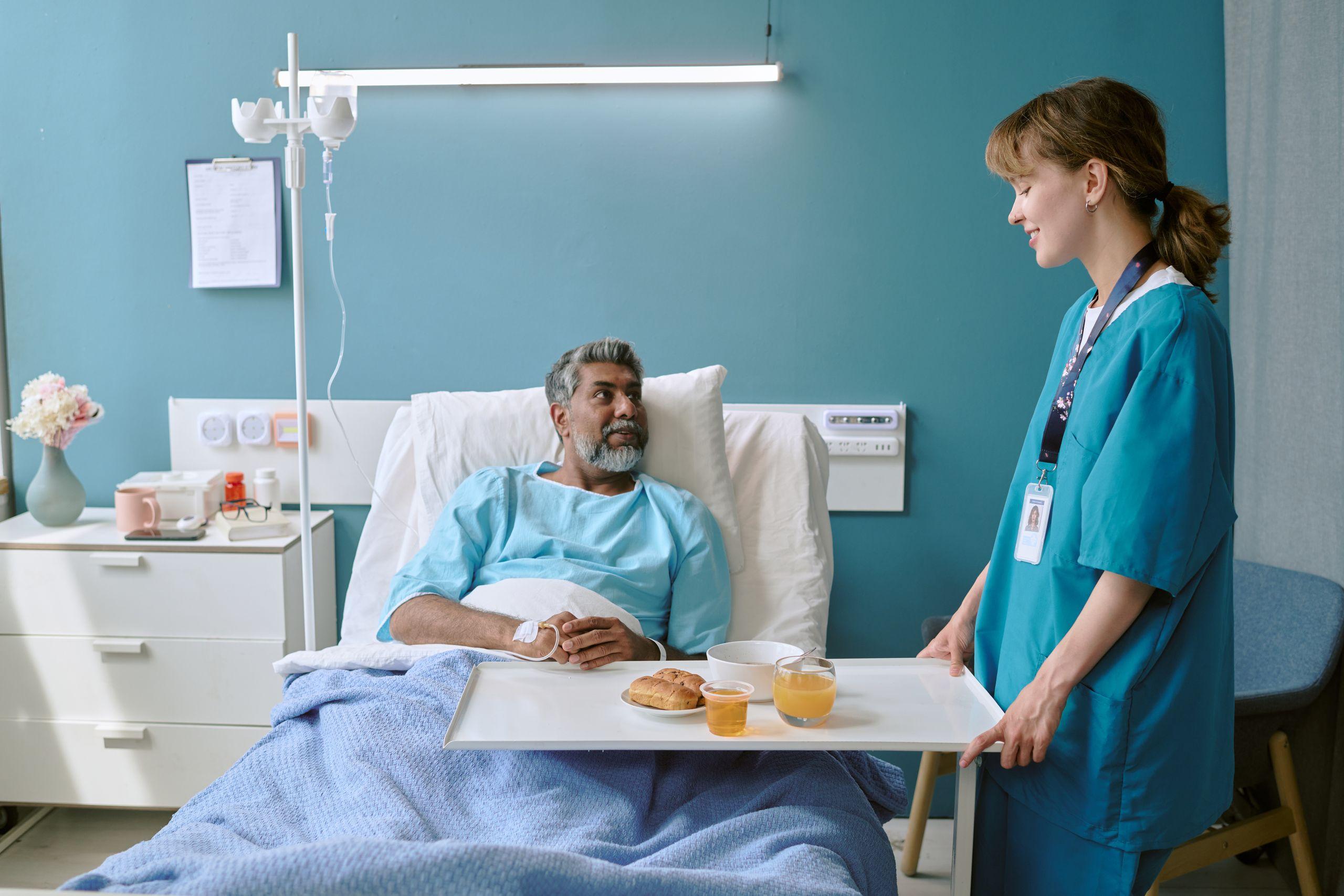 A patient in a hospital bed talks to a nurse holding a tray with food and drinks in a hospital room.