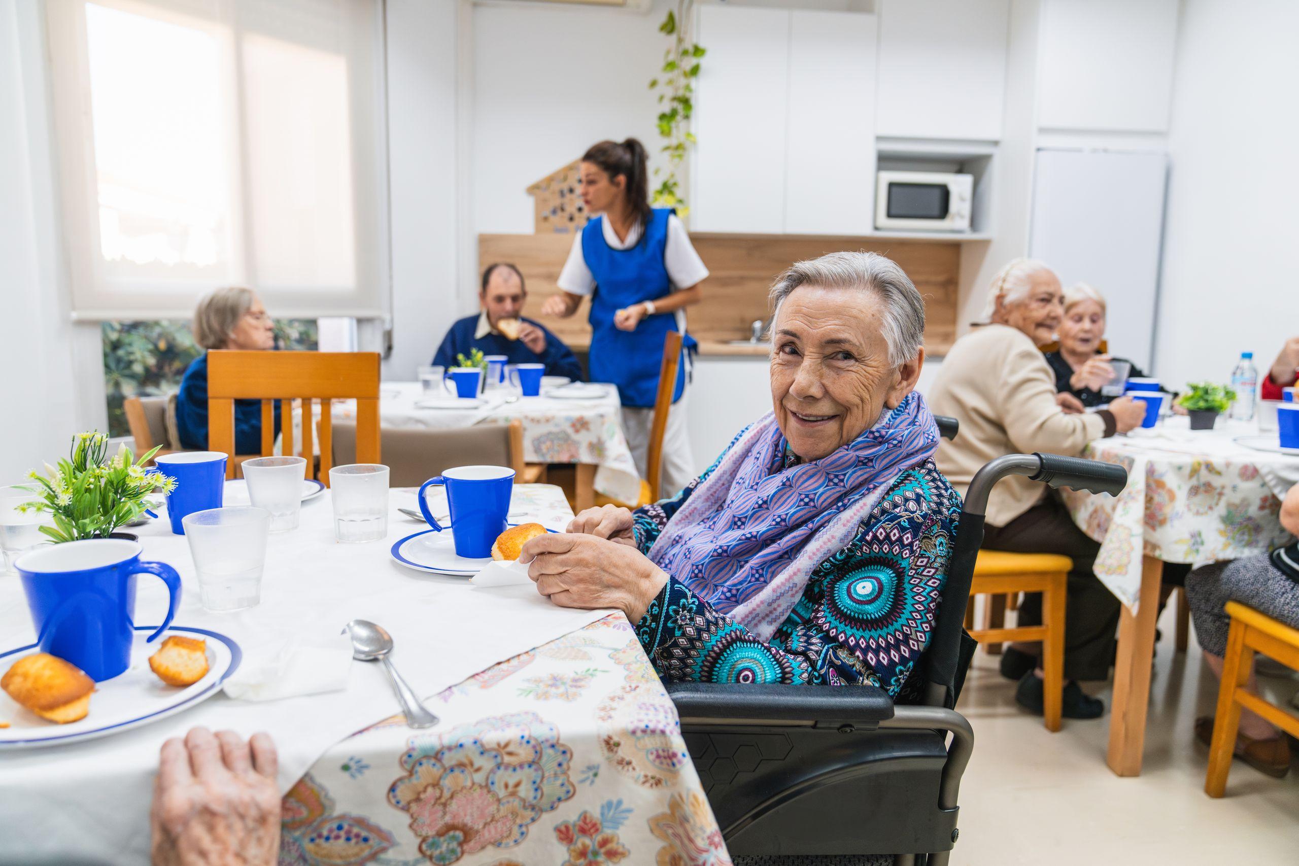 Elderly woman in a wheelchair smiles at a table with others, enjoying a meal in a bright dining area. Caregiver in the background.